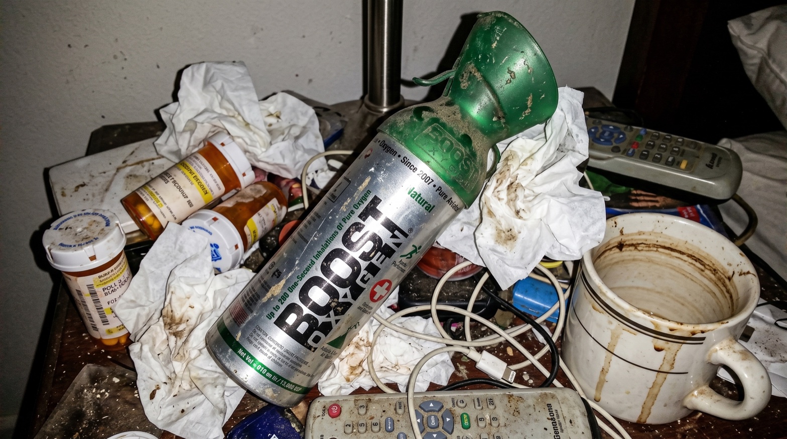 A dusty, used Boost Oxygen canister on a cluttered nightstand among old pill bottles, tissues and a stained mug
