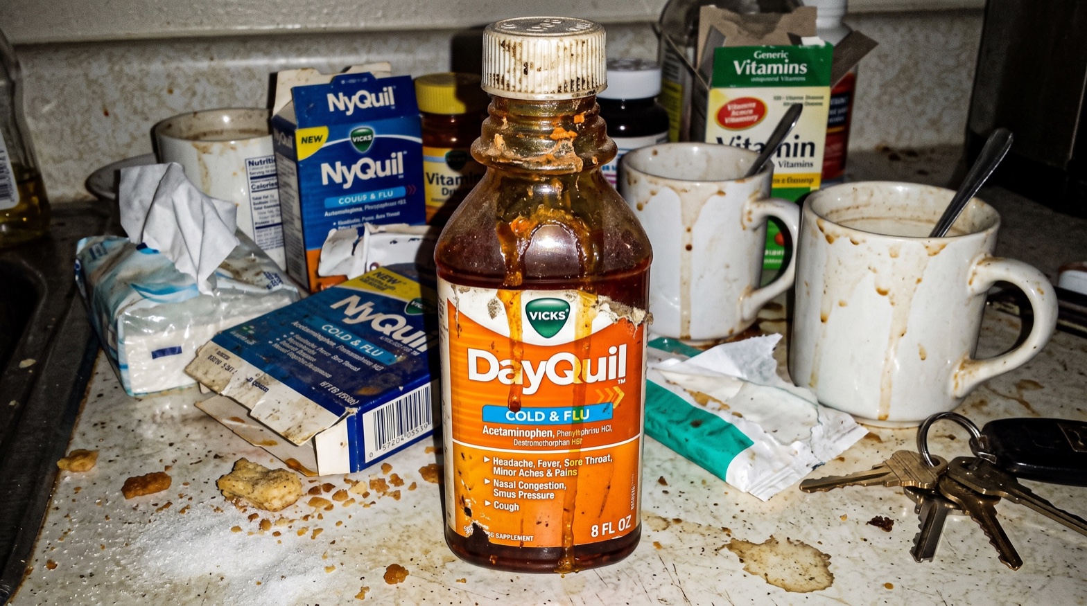 An orange Vicks DayQuil bottle on a kitchen counter in daytime, beside a steaming coffee mug and car keys