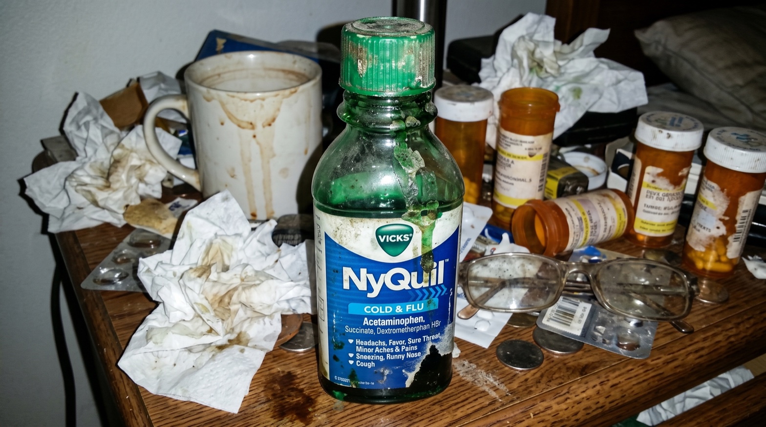 A green Vicks NyQuil bottle glowing on a nightstand at night, beside a glass of water and a used tissue
